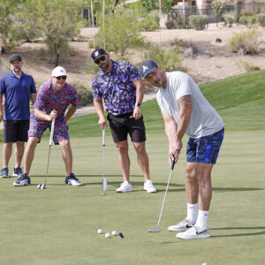 Four men in colorful outfits play mini-golf on a sunny day, with one aiming to putt on the green.