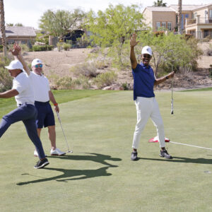 Three men on a golf course celebrate, two waving and one jumping with joy, on a sunny day.