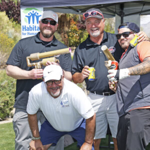 Four men pose and smile at a golf event, holding drinks and props, with a Habitat for Humanity sign behind them.