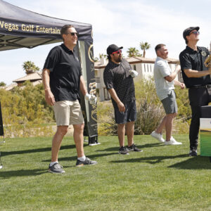 Four men stand on grass at a golf event; one aims a golf ball launcher, others watch and smile.