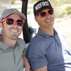 Two men wearing red sunglasses and hats smile while sitting in a golf cart on a sunny day.