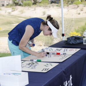 A woman in golf attire signs a board on a table outdoors at a golf course event.