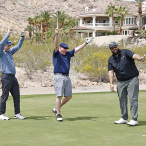 Three men celebrate on a golf course green, with two raising arms and one smiling, holding golf clubs.
