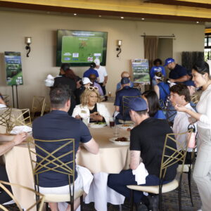 People sit at round tables eating and talking at an indoor event with a golf theme and screens in the background.