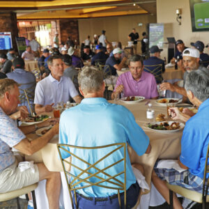 People sitting around tables eating together at a golf event in a spacious indoor venue.