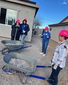 Four women in pink hard hats stand outside with wheelbarrows filled with rocks between two houses.
