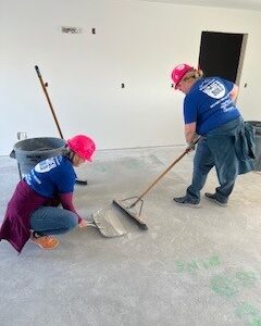 Two people in pink hard hats sweeping and cleaning a concrete floor inside a building under construction.