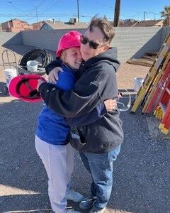 Two people in casual clothes hug outdoors at a construction site, both smiling and wearing sweatshirts.