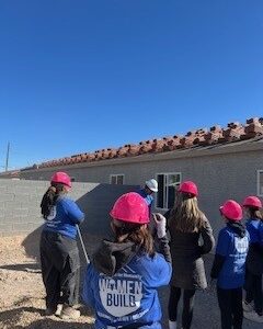 A group of women in pink hard hats and blue Women Build shirts stand outside a house under construction.