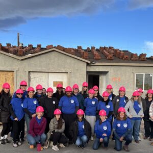 A group of people in pink hard hats pose in front of a house under construction.