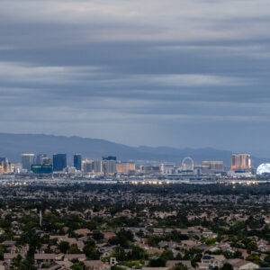 Las Vegas skyline at dusk with mountains in the background and houses in the foreground under a cloudy sky.
