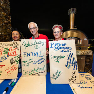 Three smiling people hold boards covered in colorful handwritten messages at an indoor event table.