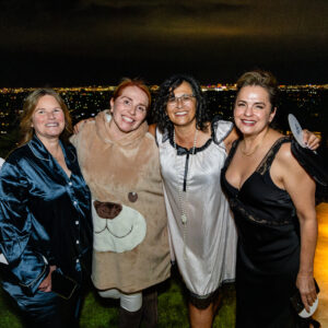 Four women posing together outdoors at night, smiling, with a city skyline glowing in the background.