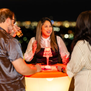Three people standing at a glowing table, laughing and talking at night with city lights in the background.