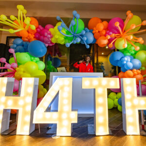 DJ behind a booth with colorful balloons and large lit-up letters spelling H4TH.