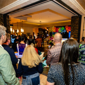 A crowd of people stands indoors, facing a festive room decorated with colorful balloons.