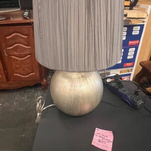 A round silver lamp with a pleated gray shade sits on a black table in a store.