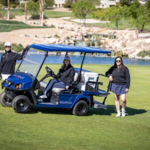 Three people posing with a blue golf cart on a golf course near a pond and a flag on a sunny day.