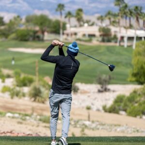 A person in a winter hat swings a golf club on a sunny golf course with mountains in the background.