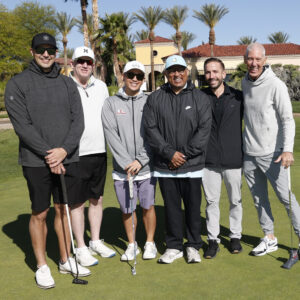 Six men stand together on a golf course, posing with golf clubs and smiling on a sunny day.