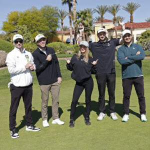 Five people posing and smiling on a golf course, holding golf clubs, with palm trees in the background.