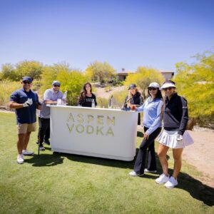 Six people stand by an Aspen Vodka bar outdoors on a sunny golf course, posing and smiling.