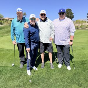 Four men with golf clubs stand together on a golf course, smiling under a clear blue sky.