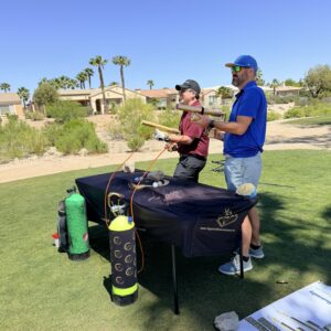 Two men aim a golf ball launcher at a golf course, with tanks and a table in front of them on a sunny day.