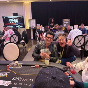 Two people pose and smile at a poker table, holding cards and making playful hand gestures.