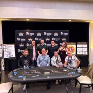 Eight people pose around a poker table with chips and cards, in front of event sponsor banners.