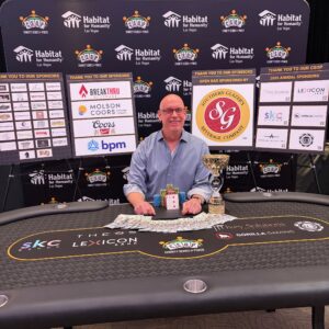 A smiling man sits at a poker table with chips, cards, and a trophy, in front of sponsor banners.