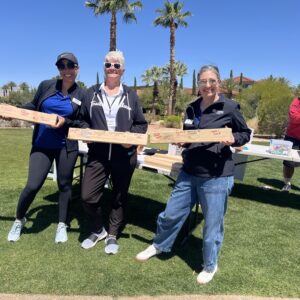 Three women smiling and holding wooden plaques outdoors on a sunny day with palm trees in the background.