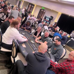 People play poker around a large table in a busy casino room with a digital clock in the background.