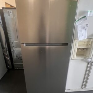 A stainless steel top-freezer refrigerator stands next to a white refrigerator in a warehouse setting.