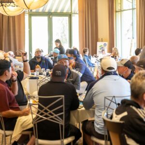 People seated at round tables in a bright room, socializing and eating during a casual event.