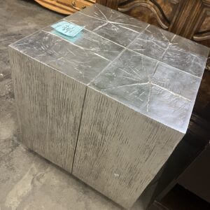 A metallic silver cubic side table with a textured surface, placed on a concrete floor near wooden furniture.