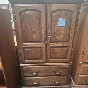 Tall wooden armoire with double doors, brass handles, and two drawers at the bottom, displayed in a store.