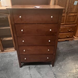 A tall wooden dresser with five drawers and silver knobs, placed on a concrete floor in a furniture store.