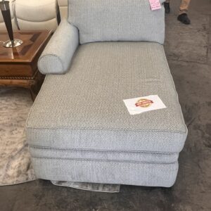 A gray upholstered chaise lounge with a bolster pillow, next to a wooden side table, sits on a concrete floor.