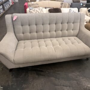 Light gray tufted mid-century modern sofa with angled arms in a furniture store setting.