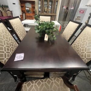 Dark wood dining table with six patterned chairs and a green leafy centerpiece in a showroom.