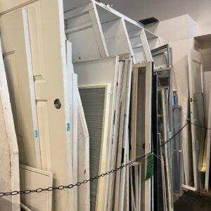 Assorted doors and window frames are stacked upright in a store behind a black chain barrier.