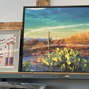Framed photo of a desert scene with cacti, next to framed architectural and abstract art, on a shelf.