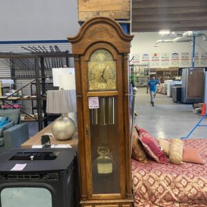 Tall wooden grandfather clock in a furniture store, with lamps and couches nearby, and a person in the background.