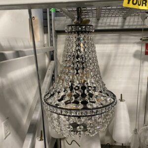 Crystal chandelier with hanging beads sits on a shelf below yellow caution tape in a storage area.