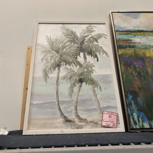 A framed painting of three palm trees by the sea sits on a shelf next to a colorful landscape painting.