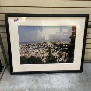 Framed photo of a cityscape with trees, displayed on a gray shelf against a slatted wall.