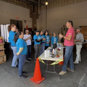 A group of people listen to a man giving instructions in a warehouse near a table with supplies.