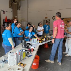 A group of people in a workshop listens to an instructor near a table with tools and cleaning supplies.