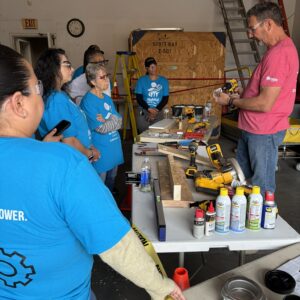 A man demonstrates power tools to a group of women in blue shirts in a workshop setting.